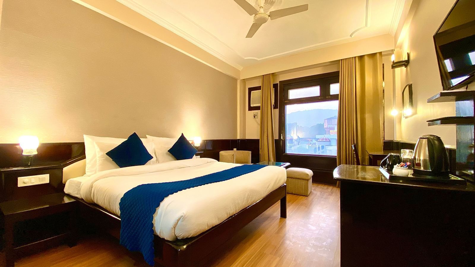 A hotel room with a king size bed, a desk, a chair, a flat-screen TV, and a window with a view of a lake  | DLS Park Grace, Haridwar